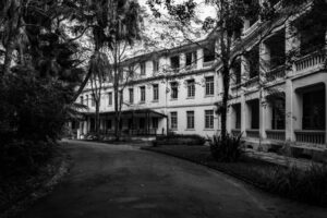 Fachada de um grande edifício histórico de três andares em preto e branco, cercado por árvores e palmeiras