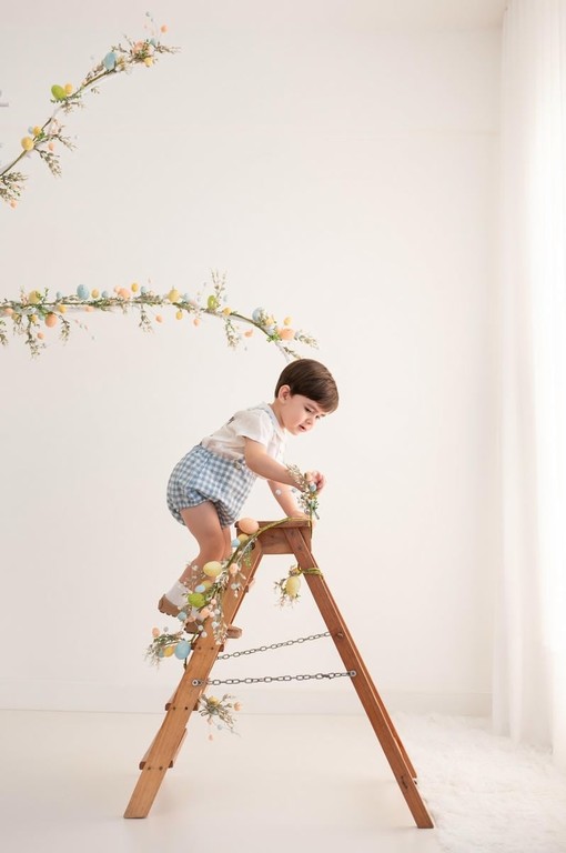 Menino pequeno subindo em escada de madeira decorada com ovos de Páscoa e flores em cenário branco
