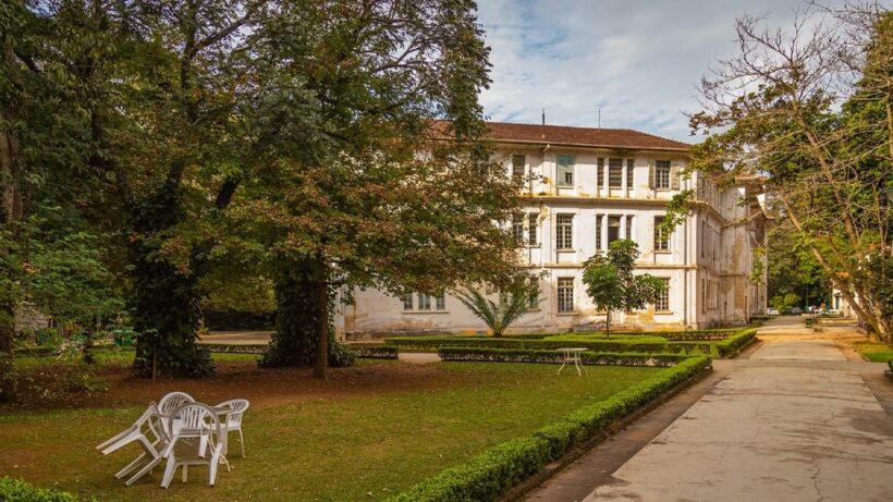 Edifício antigo de três andares com fachada branca e telhado de barro, cercado por árvores e gramado com cadeiras brancas