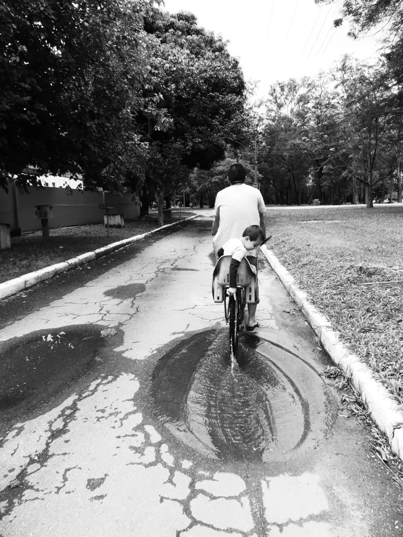 Homem e criança em uma bicicleta passando por uma poça d'água em um caminho arborizado