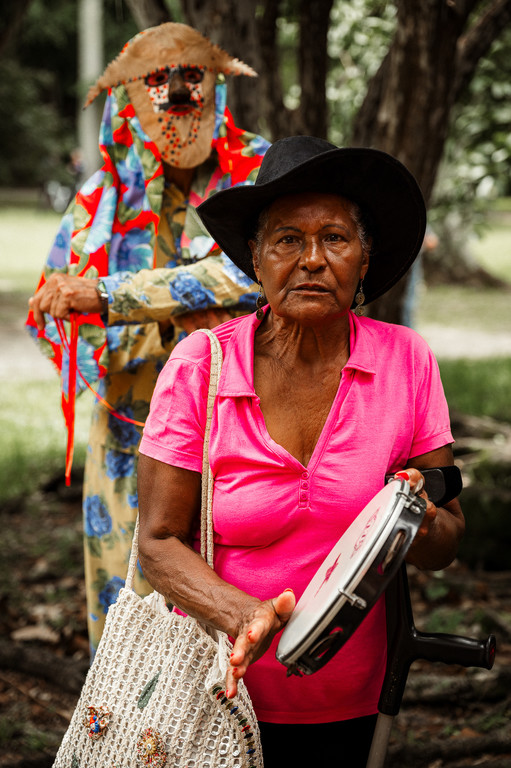 Mulher idosa de chapéu preto e blusa rosa segura um pandeiro. Ao fundo, uma figura mascarada com traje floral colorido