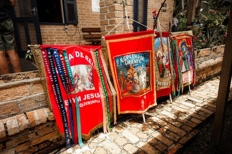 Estandartes coloridos da Folia de Reis com imagens religiosas e fitas, encostados em uma parede de tijolos