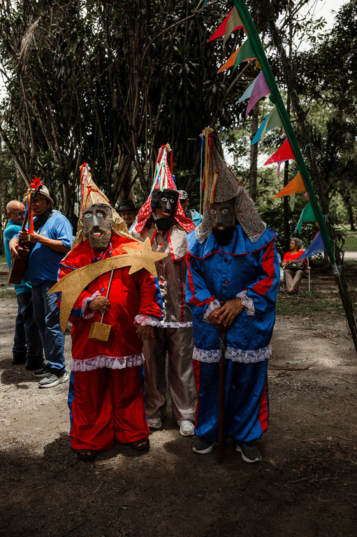 Três foliões da Folia de Reis com trajes coloridos, máscaras e chapéus cônicos; um deles segura uma estrela dourada