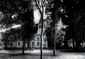 Igreja em preto e branco vista por entre árvores, com uma mulher e uma criança caminhando no pátio à frente