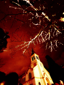Vista em ângulo baixo de uma igreja iluminada sob um céu escuro, emoldurada por galhos de árvores brilhantes