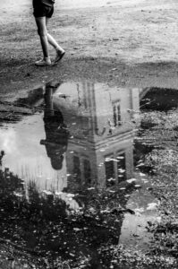Pernas de uma pessoa caminhando ao lado de uma poça que reflete sua silhueta e um edifício