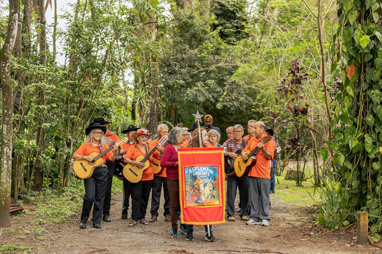Grupo musical Esplendor do Oriente tocando violões em uma trilha arborizada, com integrantes vestindo camisas laranjas