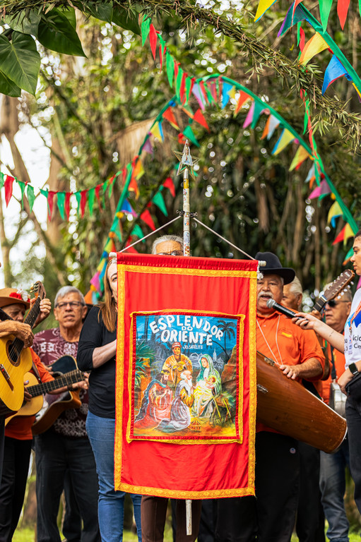 Músicos de Folia de Reis com instrumentos e estandarte vermelho escrito Esplendor do Oriente sob bandeirinhas coloridas