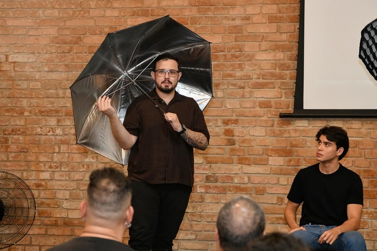 Homem segura guarda-chuva refletor de fotografia durante palestra em sala com parede de tijolos