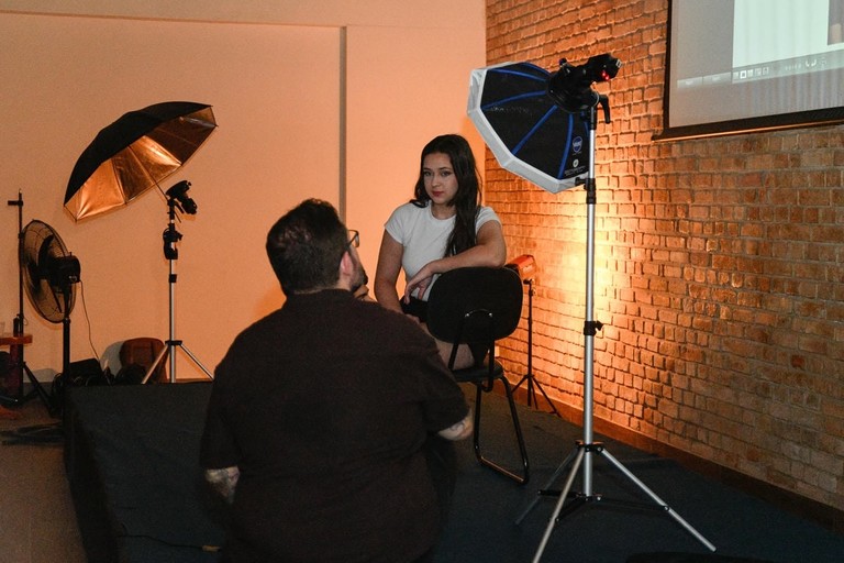Fotógrafo de costas orientando modelo em estúdio com equipamentos de iluminação profissional e parede de tijolos