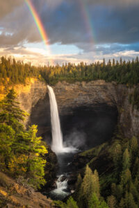 Cachoeira alta desaguando em um cânion rochoso sob um arco-íris duplo e céu nublado ao entardecer