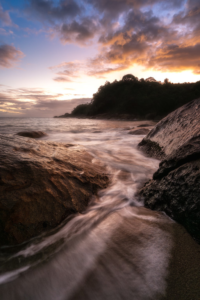 Ondas fluindo entre rochas em uma praia ao pôr do sol, com céu nublado em tons de laranja e rosa