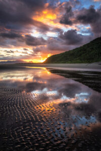 Pôr do sol em uma praia com nuvens refletidas na areia molhada e ondulada, ao lado de um morro com mata densa