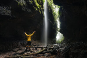 Pessoa de jaqueta amarela com braços abertos diante de uma cachoeira em uma caverna rochosa