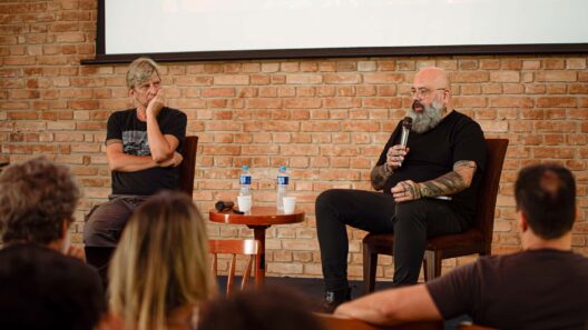 Dois homens em um painel de discussão; um fala ao microfone enquanto o outro escuta, diante de uma plateia