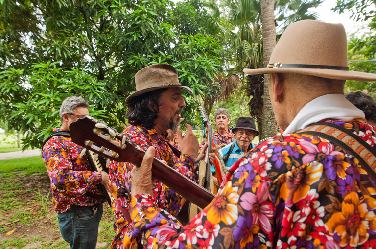 Grupo de músicos com camisas floridas e chapéus tocando violão e sanfona em um ambiente arborizado