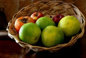 Cesta de palha com laranjas verdes, um caqui e maçãs vermelhas