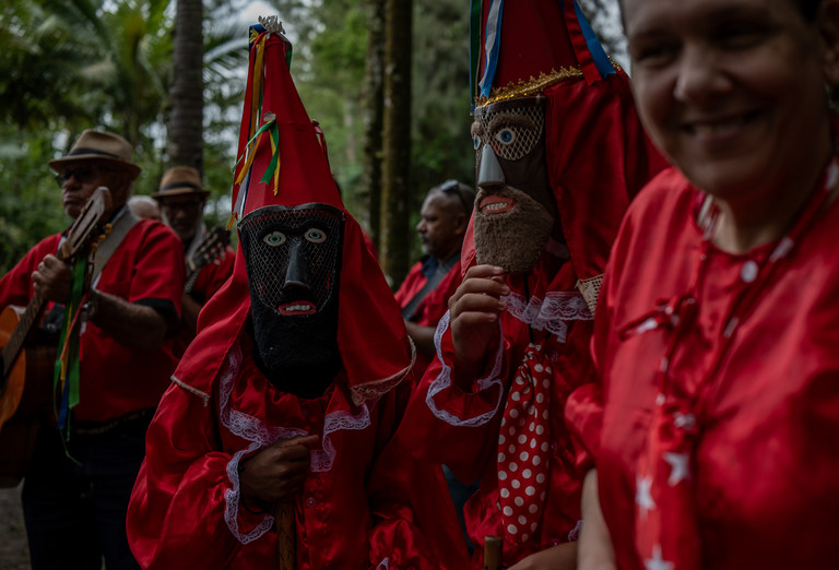 Integrantes de uma Folia de Reis com máscaras e trajes vermelhos tradicionais, acompanhados por músicos ao fundo