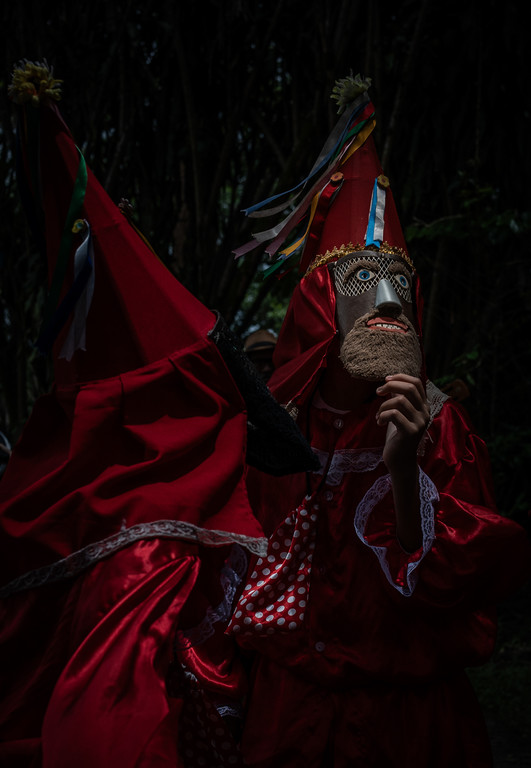 Pessoas com trajes de Folia de Reis, vestindo roupas vermelhas, chapéus cônicos e máscara artesanal com barba