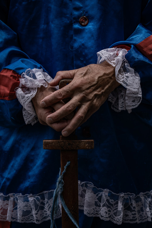 Mãos apoiadas no punho de uma espada de madeira, vestindo um traje azul com detalhes em renda branca