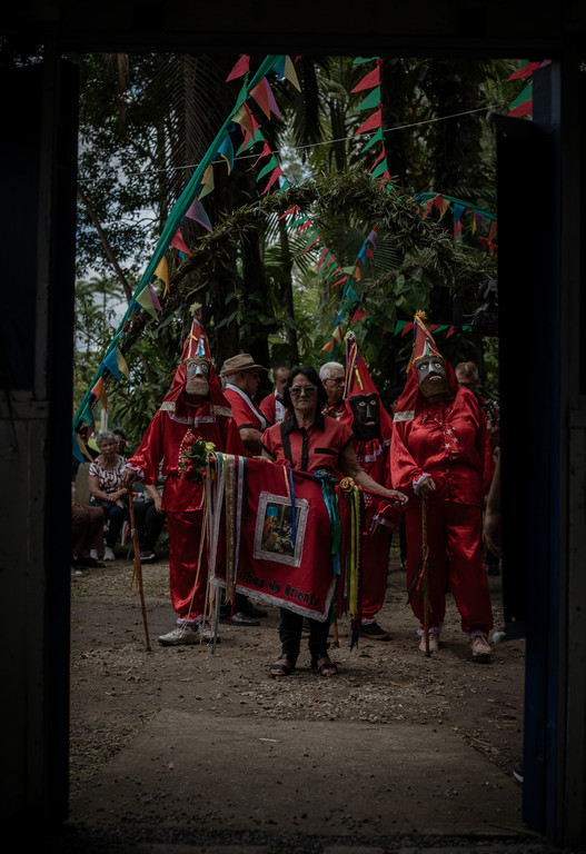 Mulher segura estandarte "Filhos do Oriente" entre palhaços de Folia de Reis mascarados sob arco de folhas e bandeirinhas