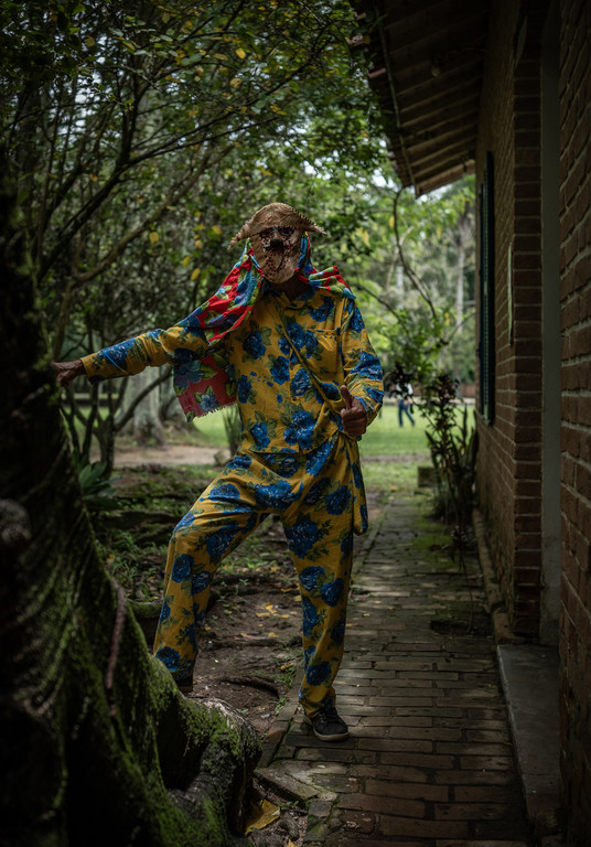 Pessoa com máscara de palha e roupa amarela florida faz sinal de positivo em um caminho de tijolos entre árvores