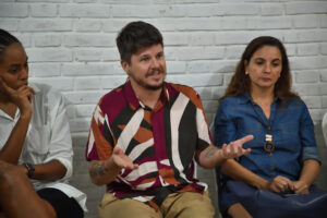 Homem de camisa estampada fala e gesticula, ladeado por duas mulheres que o escutam, em frente a uma parede de tijolos