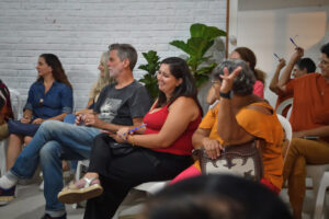 Pessoas sentadas em um evento, sorrindo e participando, em uma sala com parede de tijolos brancos