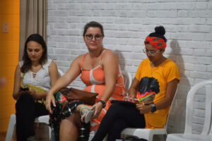 Três mulheres sentadas em cadeiras brancas contra uma parede de tijolos. Uma olha o celular e outra usa um tablet