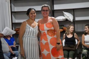 Duas mulheres sorridentes posam juntas em um ambiente interno; uma usa vestido listrado e a outra vestido com estampa laranja