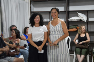 Duas mulheres sorridentes em pé no centro de uma sala com outras pessoas sentadas ao fundo