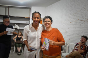 Duas mulheres sorridentes em um evento; uma veste camisa branca e a outra, de camiseta laranja, segura um brinde