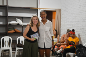 Duas mulheres sorrindo em um evento; uma mulher branca de regata preta e uma mulher negra de camisa branca