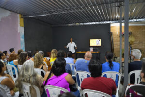 Mulher de camisa branca gesticula durante palestra para público sentado em cadeiras brancas
