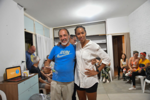 Homem de camiseta azul e mulher de camisa branca posam sorridentes em um evento com outras pessoas ao fundo