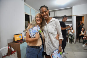Duas mulheres sorridentes se abraçam e seguram pequenos brindes em um evento interno