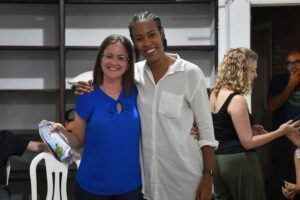 Duas mulheres sorridentes abraçadas; uma veste blusa azul e segura um pequeno kit, a outra veste camisa branca