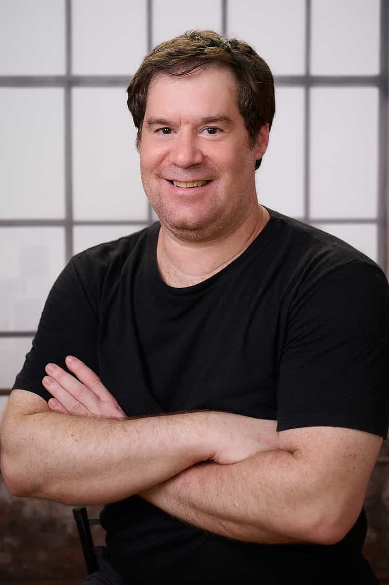 Retrato de Paulo Barbuto: homem de camiseta preta, braços cruzados, sorrindo, diante de uma parede de vidro industrial.