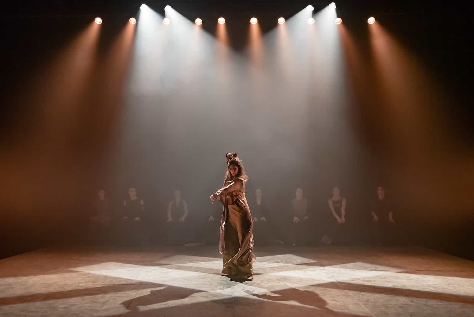 Palco de teatro atravessado por feixes de refletores em contraluz; ao centro, uma artista sozinha em traje longo claro.