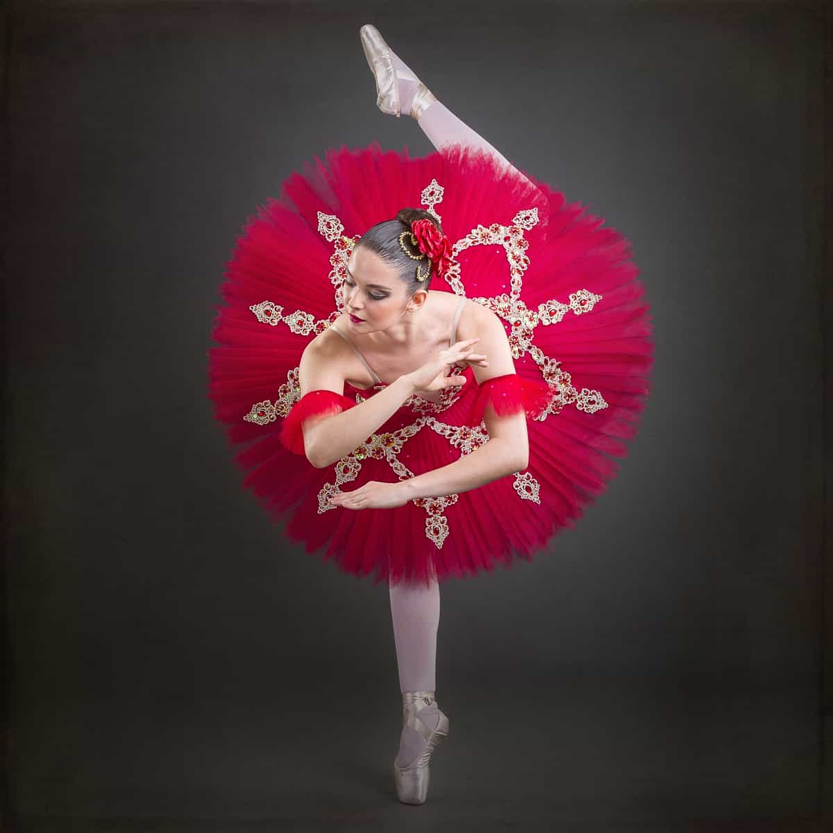 Bailarina clássica em tutu prato vermelho, sustentada na ponta com a perna esquerda em extensão vertical, sobre fundo escuro de estúdio.