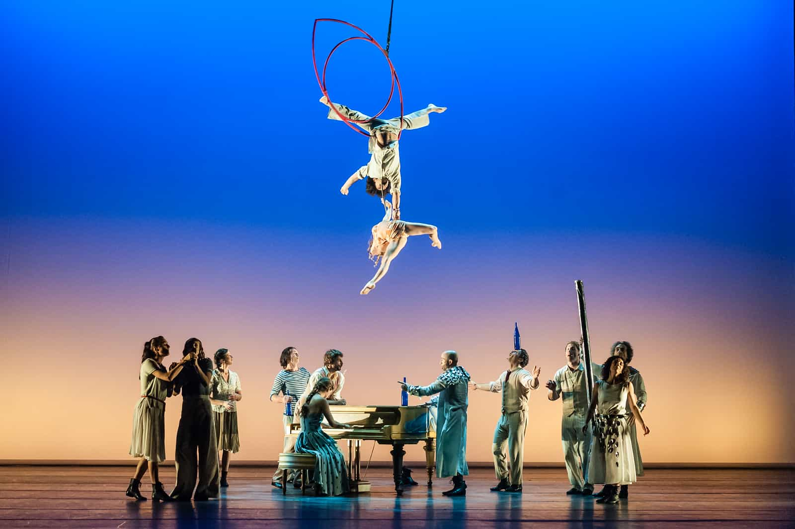 Número circense: uma acrobata suspensa no ar, de cabeça para baixo, contra um ciclorama azul intenso, com um grupo de artistas de figurino verde no palco abaixo.